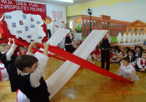 Na tle dekoracji z mapą Polski i dziećmi z chorągiewkami przedszkolaki wykonują taniec z szarfami.